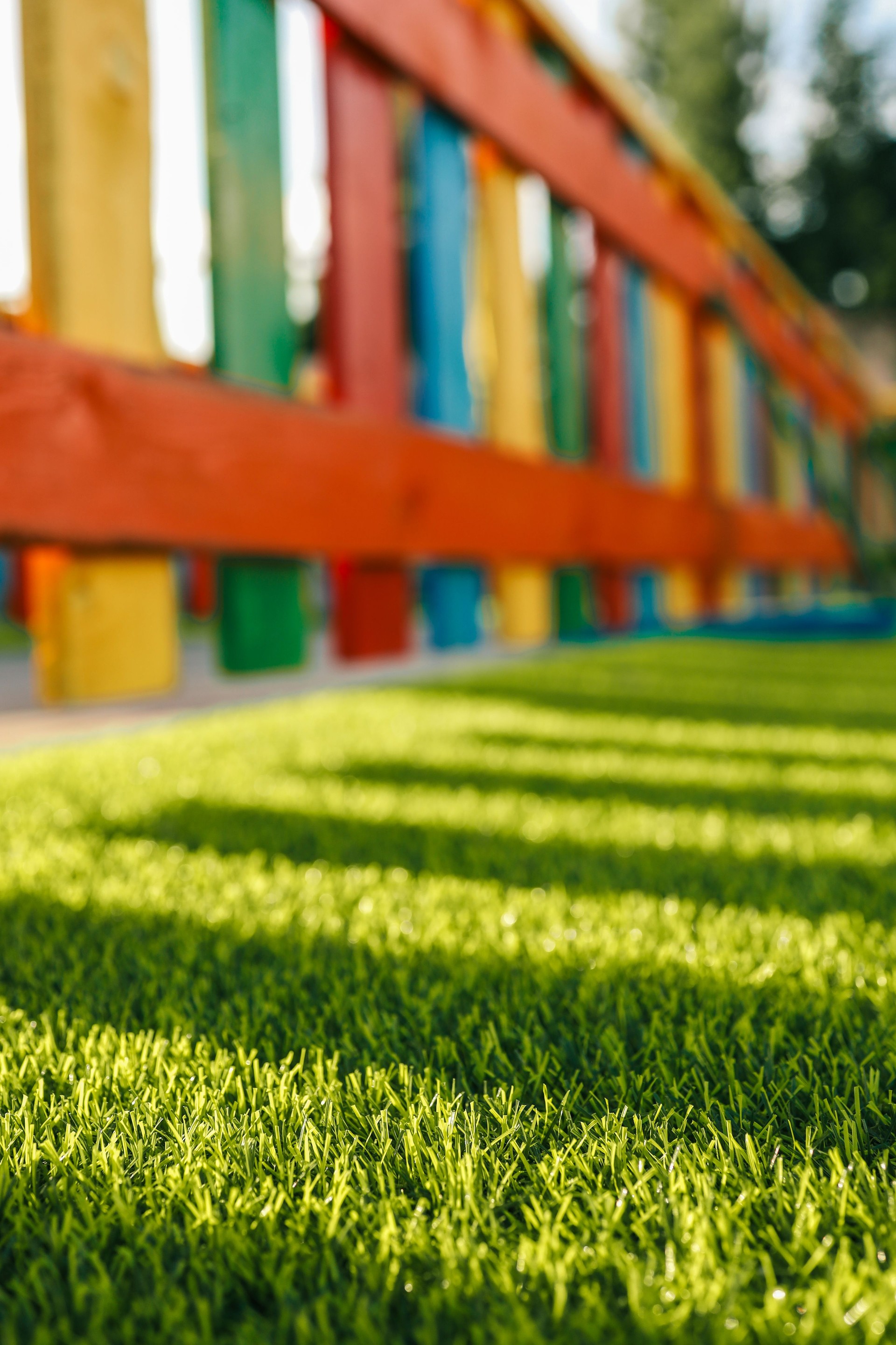 Shadows of a colorful wooden fence falling on green artificial grass in a children's playground on a sunny day. Bright, playful, and geometric composition